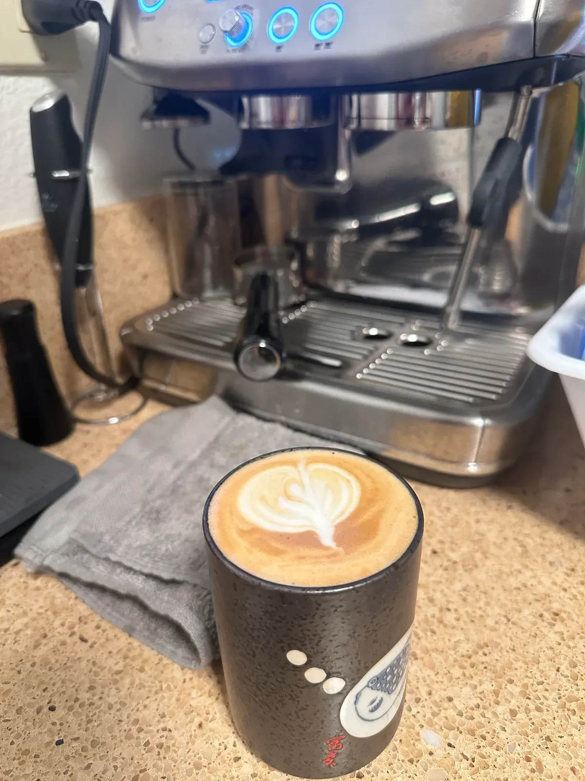 A latte with leaf art in a ceramic cup next to a home espresso machine on a kitchen counter