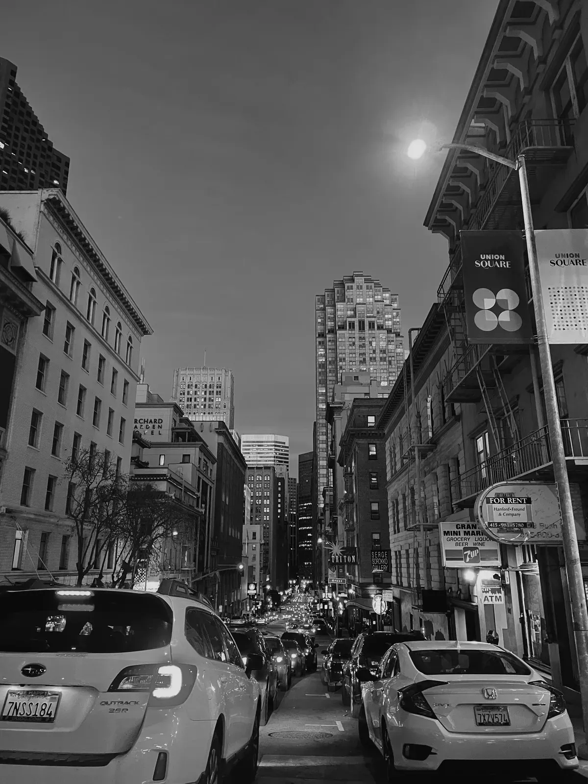 Black-and-white street photograph of a San Francisco street at dusk near Union Square, parked cars and tall buildings receding into the distance